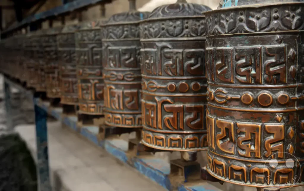 Eine Reihe von kunstvoll verzierten Gebetsmühlen aus Bronze ist in einer Reihe aufgestellt. Die Gravuren der tibetischen Schrift sind deutlich erkennbar und verleihen den Mühlen eine tiefe spirituelle Bedeutung. Die leichte Unschärfe im Hintergrund und die erdigen Töne des Metalls schaffen eine ruhige und besinnliche Atmosphäre.