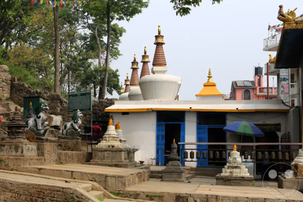Bunte Gebetsfahnen wehen über einem Tempelkomplex in Kathmandu, Nepal. Mehrere weiße Stupas mit goldenen Spitzen reihen sich entlang eines Gebäudes, das mit blauen Metallgittern geschmückt ist. Am Boden sind steinerne Skulpturen von Löwen und mehrere Reihen von Gebetsmühlen zu sehen, die eine spirituelle Atmosphäre schaffen.