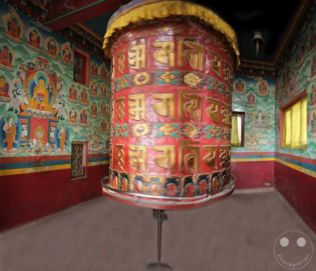 Ein großes, rot-goldenes Gebetsrad dreht sich in einem buddhistischen Kloster in Kathmandu. Die Wände sind mit farbenprächtigen Malereien verziert, die religiöse Szenen und Gottheiten darstellen. Die Atmosphäre wirkt ruhig und ehrfürchtig, trotz der Bewegung des Gebetsrades.