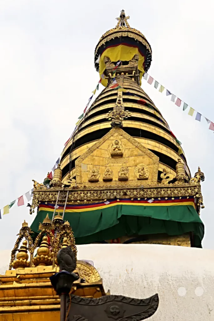 Die goldene Spitze der Swayambhunath-Stupa in Kathmandu, Nepal, ist mit bunten Gebetsfahnen geschmückt. Detailreiche Verzierungen und Buddha-Statuen zieren die Struktur, die einen Eindruck von spiritueller Erhabenheit vermittelt.