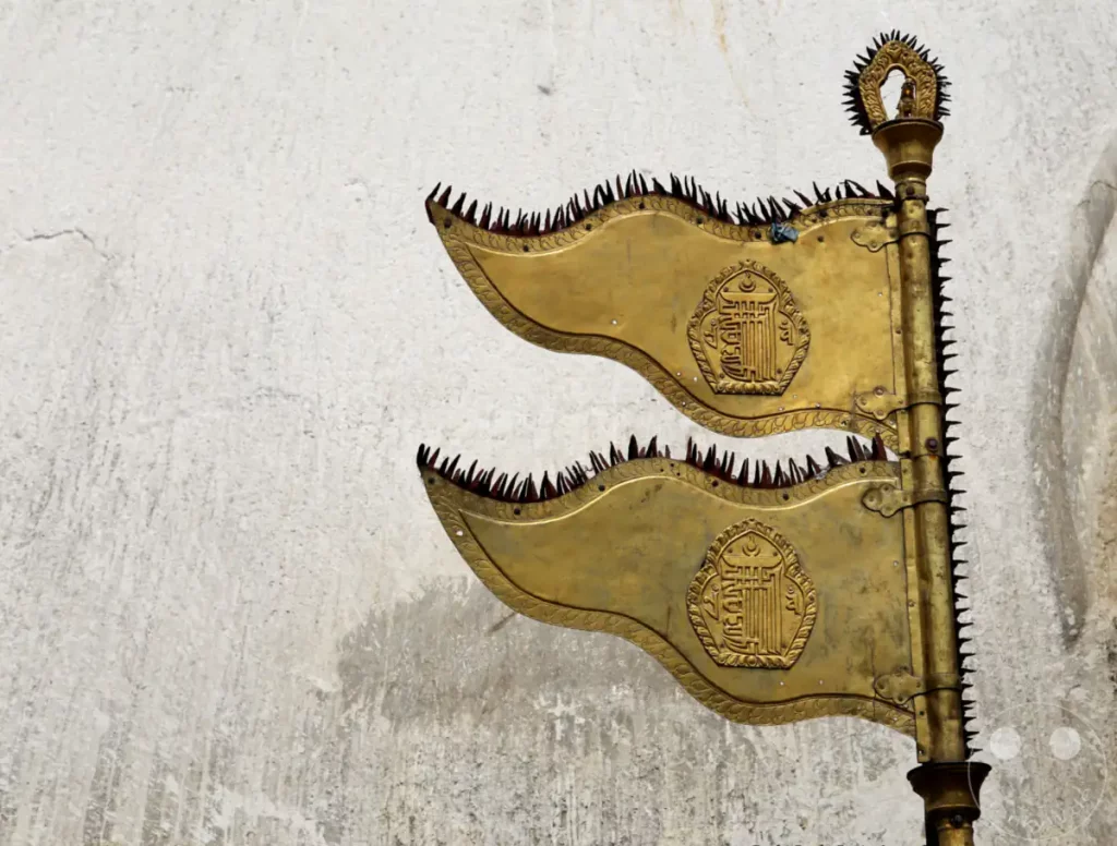 Zwei goldene, verzierte Flaggen mit spitzen Verzierungen sind an einem Mast vor der Swayambhunath-Stupa in Kathmandu, Nepal, befestigt. Die kunstvollen buddhistischen Symbole auf den Flaggen deuten auf ihre spirituelle Bedeutung hin, während die raue Textur der Wand im Hintergrund einen starken Kontrast bildet.