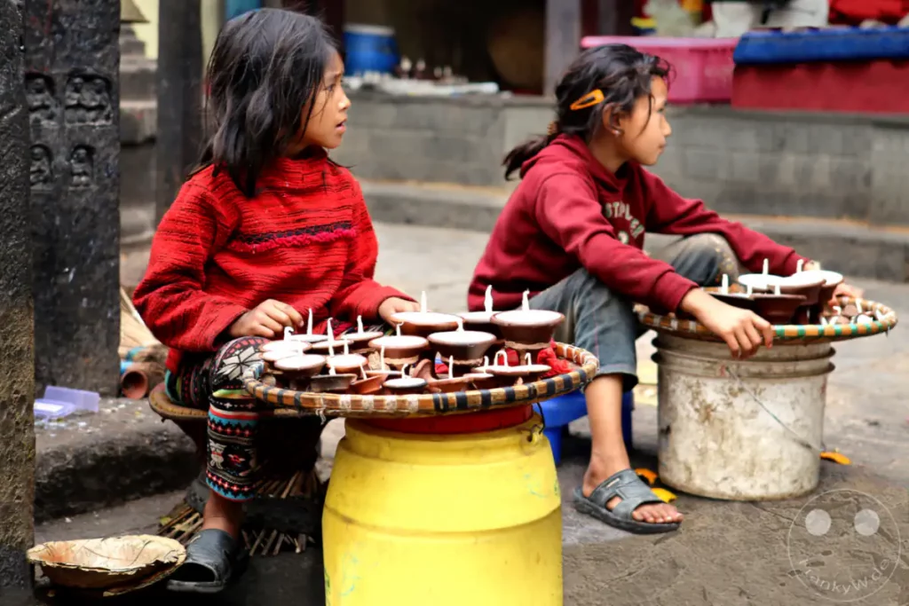 Zwei junge Mädchen sitzen in der Nähe der Swayambhunath-Stupa in Kathmandu und verkaufen kleine Tonlampen. Die Szene fängt einen Moment des Alltagslebens inmitten eines spirituellen Ortes ein und vermittelt eine ruhige, aber geschäftige Atmosphäre.
