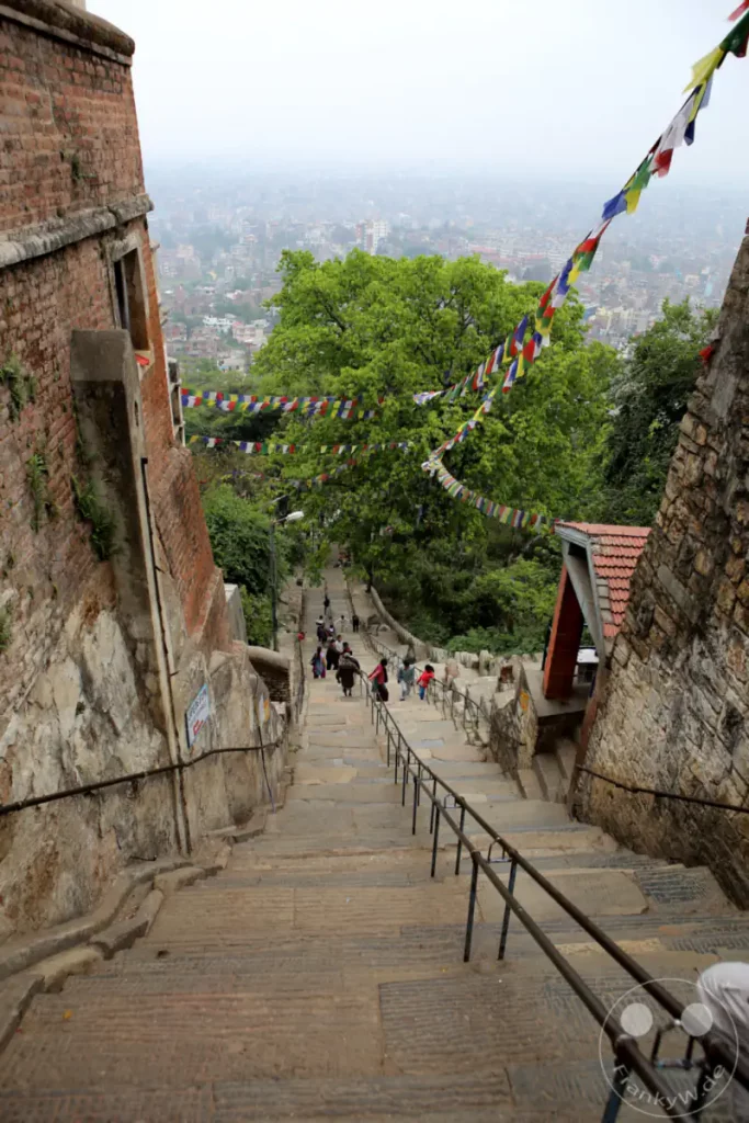 Eine lange Steintreppe führt hinunter zu einer städtischen Landschaft, die von dicht stehenden Gebäuden und dem leichten Dunst des Morgens geprägt ist. An einem Geländer entlang wehen bunte Gebetsfahnen im Wind und schaffen eine spirituelle Atmosphäre. Menschen gehen die Treppen hinauf und hinunter und beleben die Szene mit ihrer Anwesenheit.