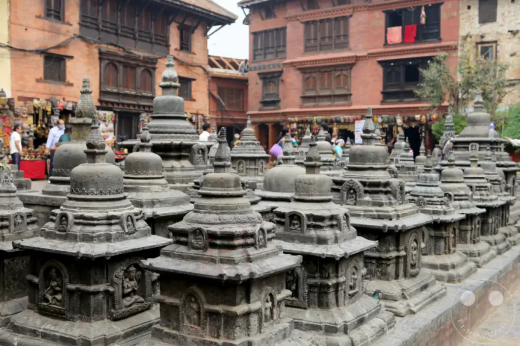 Zahlreiche kleine steinerne Stupas sind in einer Reihe aufgestellt, umgeben von traditioneller nepalischer Architektur. Die Szene strahlt eine friedliche und kulturell reiche Atmosphäre aus.