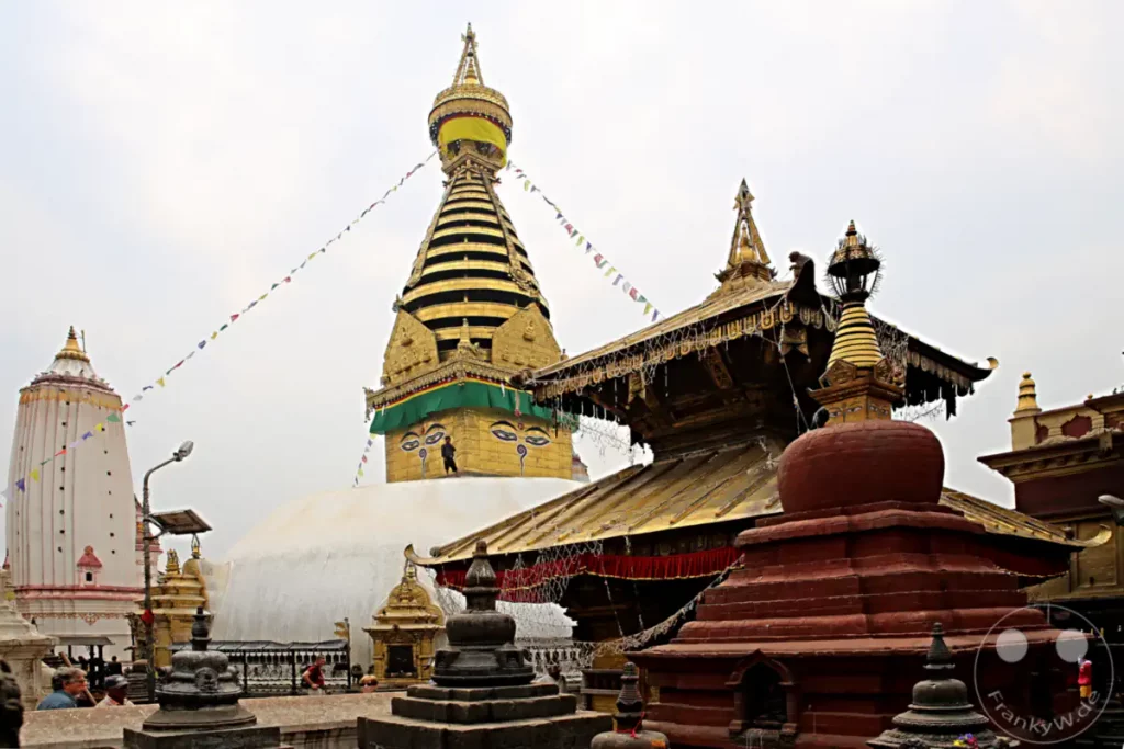 Der Swayambhunath-Tempelkomplex in Kathmandu, Nepal, ist mit goldenen Stupas, flatternden Gebetsfahnen und kunstvollen architektonischen Details geschmückt. Affen sind ein häufiger Anblick und machen die Atmosphäre des Ortes lebendig. Der Himmel ist leicht bewölkt und verleiht der Szene eine gedämpfte, aber spirituelle Stimmung.