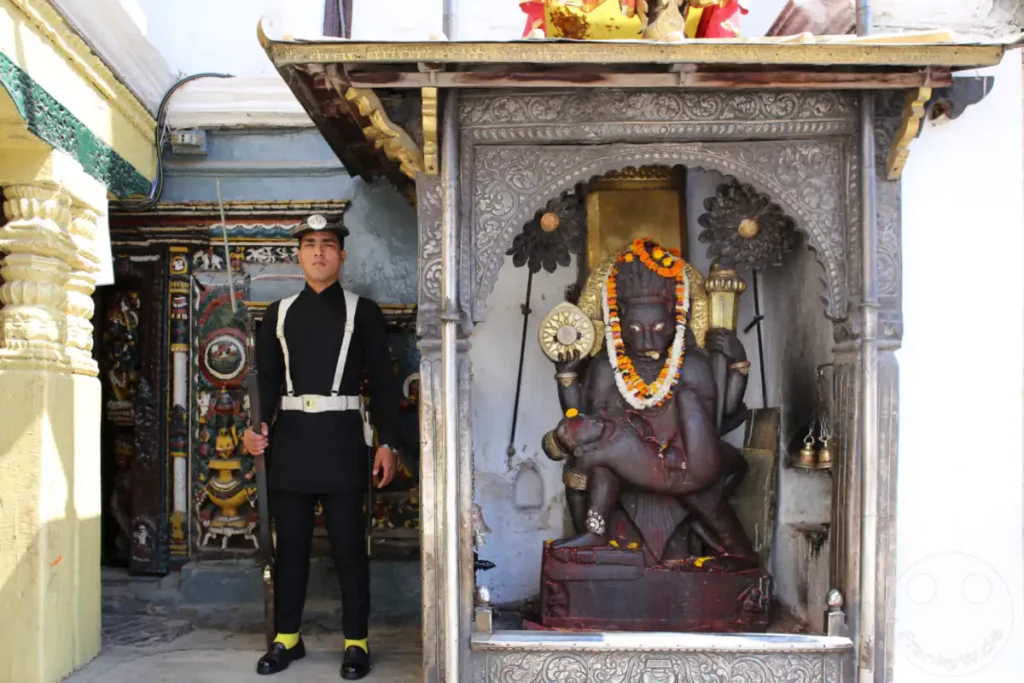 Ein Wächter in traditioneller Uniform steht vor einer aufwändig verzierten Statue eines hinduistischen Gottes in Kathmandu, Nepal. Die Szene deutet auf die religiöse und kulturelle Bedeutung dieses Ortes im Kathmandu Durbar Square hin.
