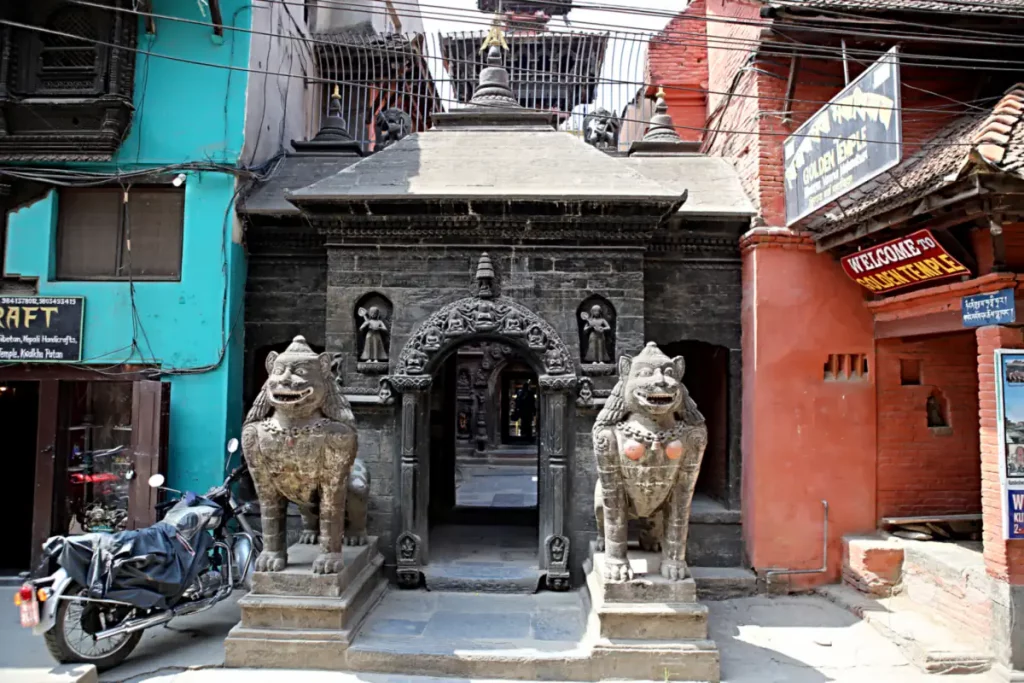 Eine traditionelle Toranlage am Hiranya Varna Mahavihar Kloster in Lalitpur, Nepal. Zwei steinerne Löwen wachen über den Eingang, flankiert von kunstvollen Schnitzereien und buddhistischen Skulpturen. Die Architektur spiegelt die reiche kulturelle und religiöse Geschichte der Region wider.