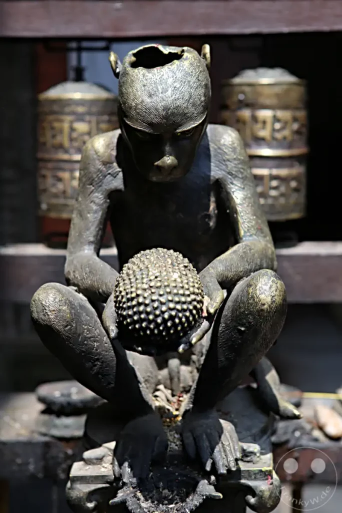 Eine detaillierte Bronzestatue eines Affen, der eine kugelförmige Frucht hält, steht im Hiranya Varna Mahavihar Kloster in Lalitpur, Nepal. Im Hintergrund sind buddhistische Gebetsmühlen zu erkennen, die zur spirituellen Atmosphäre des Ortes beitragen.