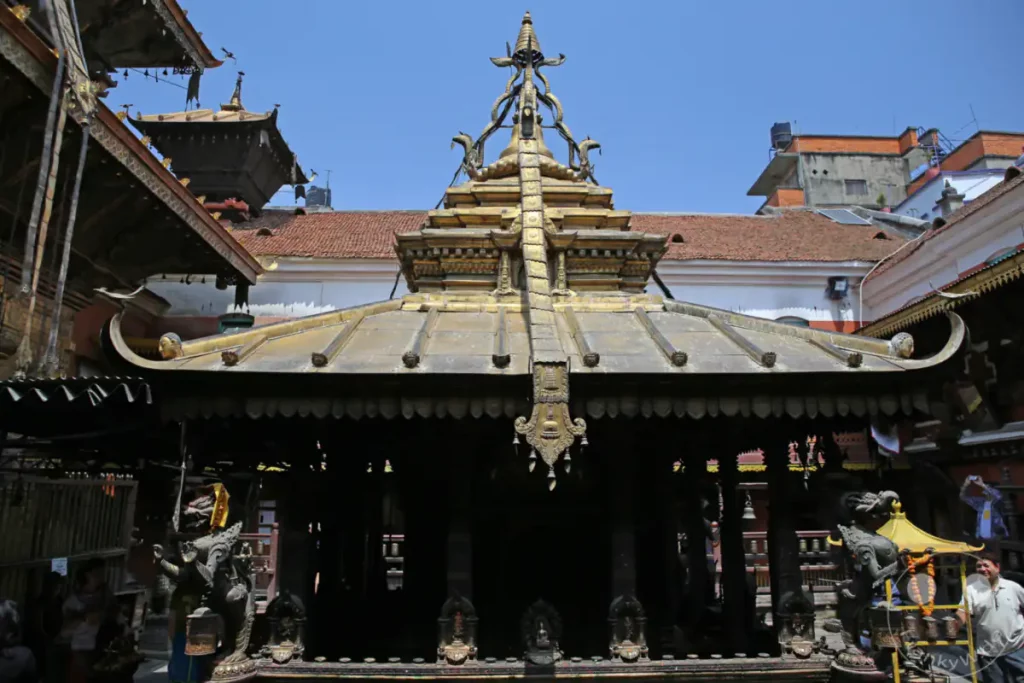 Das Bild zeigt die kunstvolle Architektur des Hiranya Varna Mahavihar Klosters in Lalitpur, Nepal. Vergoldete Dächer und filigrane Schnitzereien prägen die Szene unter einem klaren blauen Himmel. Die detailreiche Gestaltung offenbart die reiche kulturelle und religiöse Geschichte des Ortes.