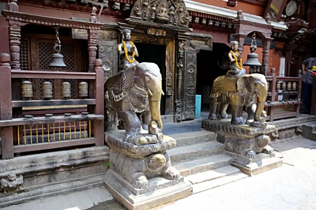 Zwei kunstvoll gestaltete Elefantenskulpturen stehen vor dem Eingang des Hiranya Varna Mahavihar Klosters in Lalitpur, Nepal. Die Skulpturen sind auf detailreichen Sockeln platziert und mit Girlanden geschmückt, was dem Ort eine feierliche Atmosphäre verleiht.