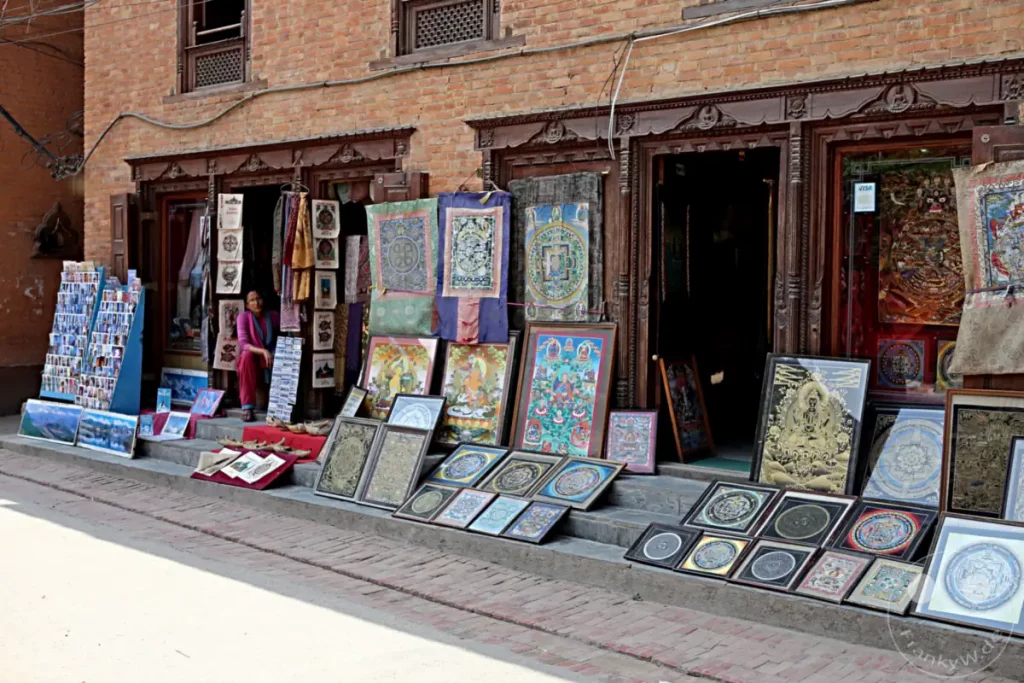 Vorderansicht eines Verkaufsstandes vor dem Hiranya Varna Mahavihar Kloster in Lalitpur, Nepal. Gerahmte Kunstwerke und Postkarten werden auf dem Gehsteig ausgestellt und verkauft, während ein Verkäufer im Hintergrund sitzt.