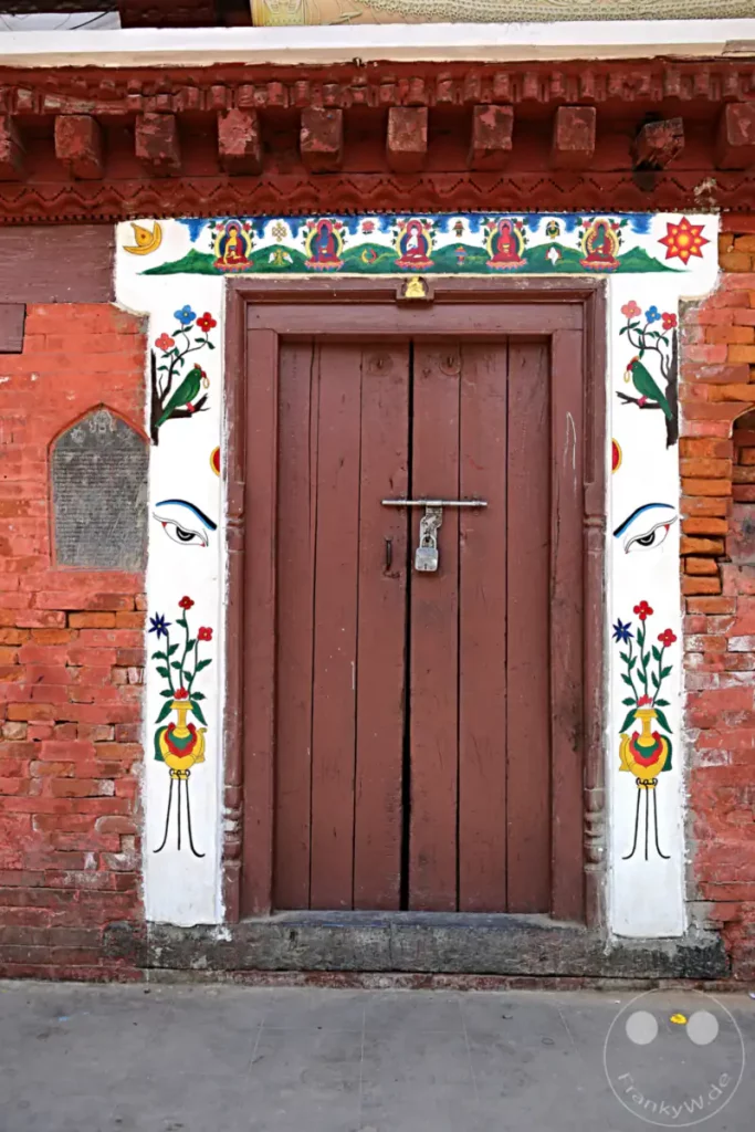 Eine traditionelle Holztür im Hiranya Varna Mahavihar Kloster in Lalitpur, Nepal. Die Tür ist mit farbenfrohen buddhistischen Motiven und Verzierungen geschmückt, die ihre kulturelle und religiöse Bedeutung unterstreichen. Die Backsteinarchitektur und die kunstvollen Malereien schaffen eine ehrwürdige Atmosphäre.
