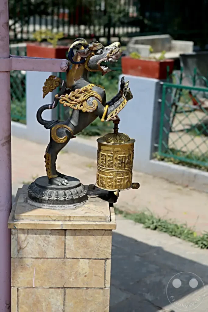 Eine kunstvolle Skulptur eines Drachenähnlichen Wesens hält eine verzierte goldene Gebetsmühle aufrecht. Die Szene spielt sich vor dem Hintergrund des buddhistischen Klosters Hiranya Varna Mahavihar in Lalitpur, Nepal, ab und strahlt eine religiöse und kulturelle Atmosphäre aus.