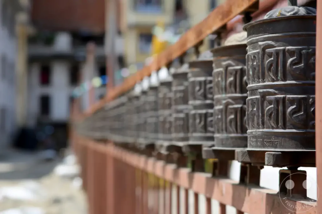 Eine Reihe von buddhistischen Gebetsmühlen ist entlang einer metallischen Geländerreihe aufgereiht, wobei die vorderen Mühlen scharf abgebildet und die hinteren unscharf sind. Die Gebetsmühlen zeigen eingravierte Schriftzeichen und sind aus dunklem Metall gefertigt, auf einem rudimentären Metallrahmen montiert. Die Aufnahme vermittelt ein Gefühl der Besinnung und spirituellen Atmosphäre, die typisch für religiöse Stätten in Kathmandu ist.