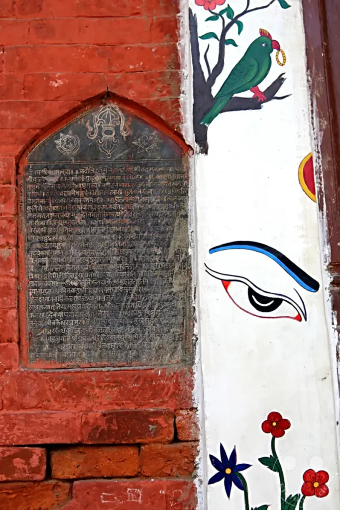 Eine detaillierte Aufnahme einer verzierten Klosterwand im Hiranya Varna Mahavihar Kloster in Lalitpur, Nepal. Die Wand zeigt eine alte Steininschrift und bunte Malereien, darunter ein Vogel und ein Auge, die eine kulturelle und künstlerische Bedeutung vermitteln.