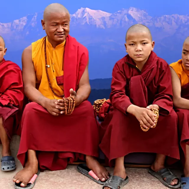 Vier junge Mönch-Schüler sitzen in ihren traditionellen roten Roben vor der Kulisse des Himalaya-Gebirge. Zwei von ihnen halten Meditationsperlen in den Händen und blicken nachdenklich in die Kamera. Die Szene strahlt eine ruhige und kontemplative Atmosphäre aus.