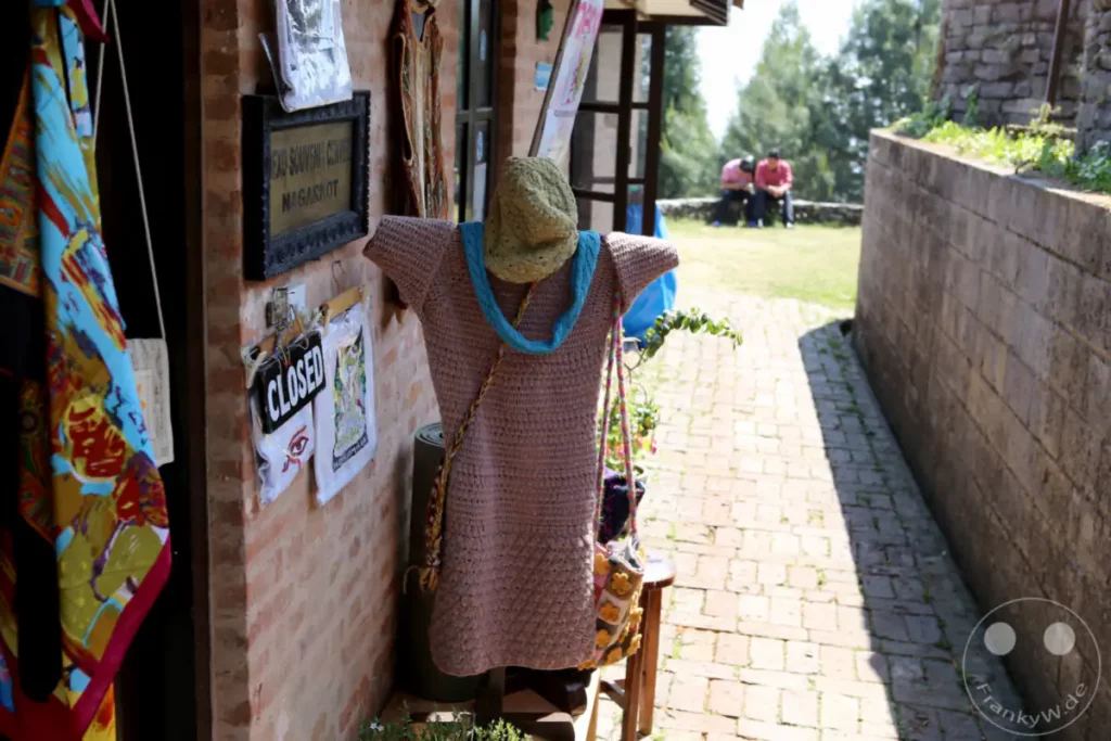 Eine Schaufensterpuppe steht vor einem Souvenir-Laden in Nagarkot, Nepal. SIe trägt eine gestrickte Tunika, einen Hut und eine Tasche. Im Hintergrund sitzen zwei Männer auf einer Bank und unterhalten sich in einer ländlichen Umgebung.
