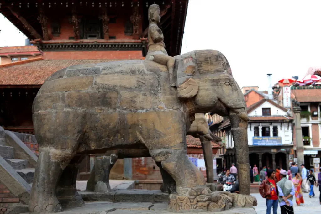 Eine detailreiche Steinstatue eines Elefanten mit einem Reiter steht im Vordergrund des Bildes am Durbar Square in Patan, Nepal. Im Hintergrund ist traditionelle nepalesische Architektur zu sehen, die die historische Bedeutung des Ortes unterstreicht. Die Szene vermittelt einen Eindruck von kulturellem Erbe und lokaler Atmosphäre.