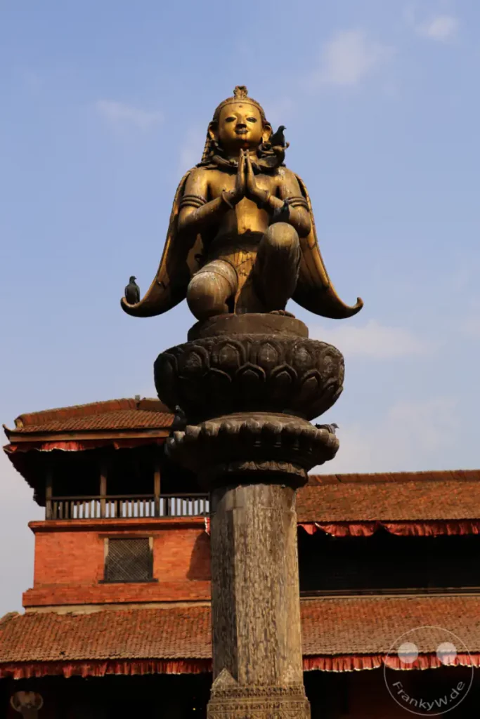 Eine goldene Garudastatue auf einem verzierten Sockel, die den Betrachter mit gefalteten Händen anspricht. Im Hintergrund sind traditionelle Ziegeltempel mit roten Dachziegeln zu sehen, und mehrere Tauben sitzen auf der Statue und dem Sockel. Das Bild vermittelt eine ruhige und kulturell reiche Atmosphäre im historischen Durbar Square von Patan.