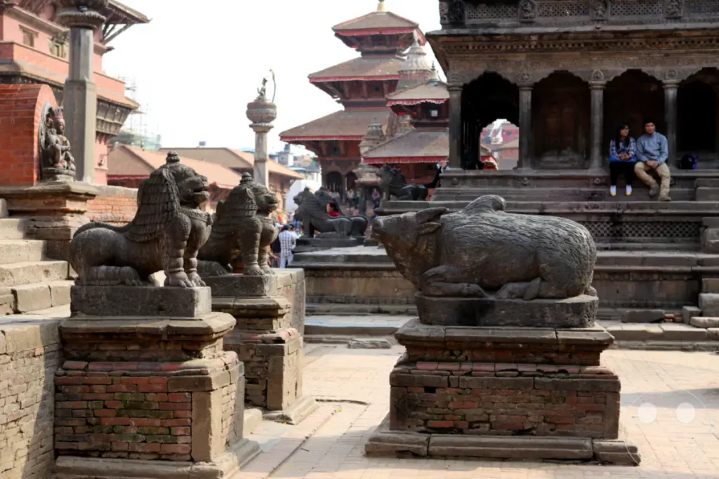 Zwei steinerne Löwen und eine Büffelstatue stehen im Vordergrund des Durbar Square in Patan, Nepal. Im Hintergrund sind traditionelle nepalesische Tempelarchitektur und einige Besucher zu sehen. Die Szene vermittelt einen Eindruck von der historischen und kulturellen Bedeutung des Ortes.