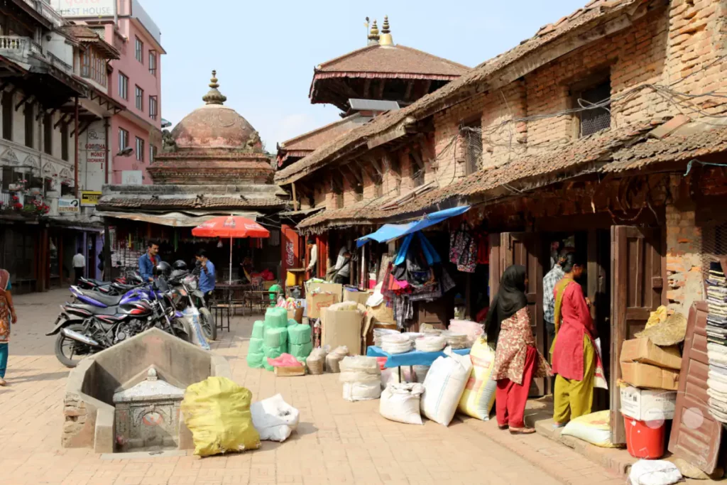 Der lebhafte Durbar Square in Patan, Nepal, ist ein geschäftiger Marktplatz, auf dem Händler Waren feilbieten. Traditionelle Gebäude säumen den Platz und schaffen eine historische Kulisse für das tägliche Treiben. Kunden und Händler interagieren inmitten von Waren, Motorrädern und architektonischen Wahrzeichen.