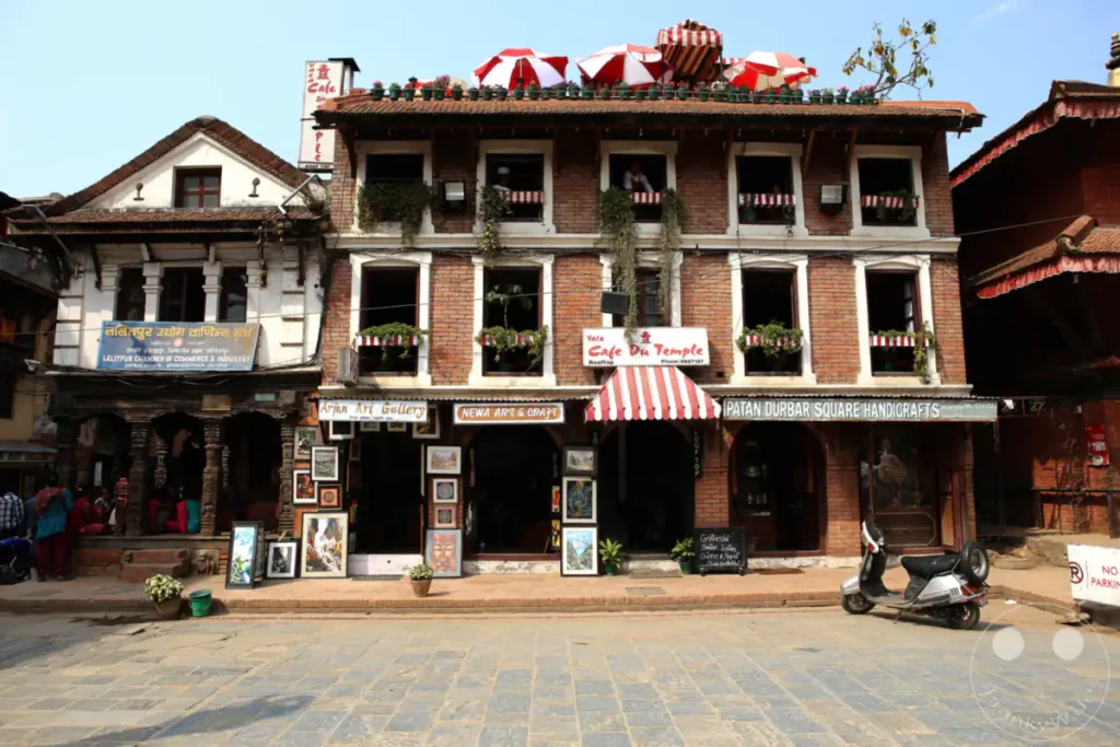 Eine belebte Straße vor kunstvollen Gebäuden am Durbar Square in Patan, Nepal. Geschäfte für Kunsthandwerk und Galerien säumen den Platz, während eine Vespa davor geparkt ist und eine entspannte Atmosphäre herrscht.