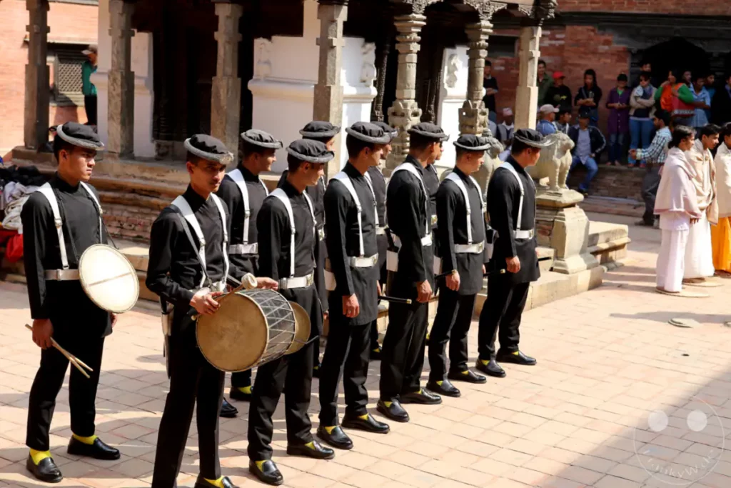 Eine Gruppe von Trommlern in traditioneller Kleidung steht in einer Reihe auf dem historischen Durbar Square in Patan, Nepal. Sie tragen schwarze Uniformen mit weißen Akzenten und schwarzen Hüten. Die Szene vermittelt eine feierliche und kulturell reiche Atmosphäre während eines traditionellen Festes.