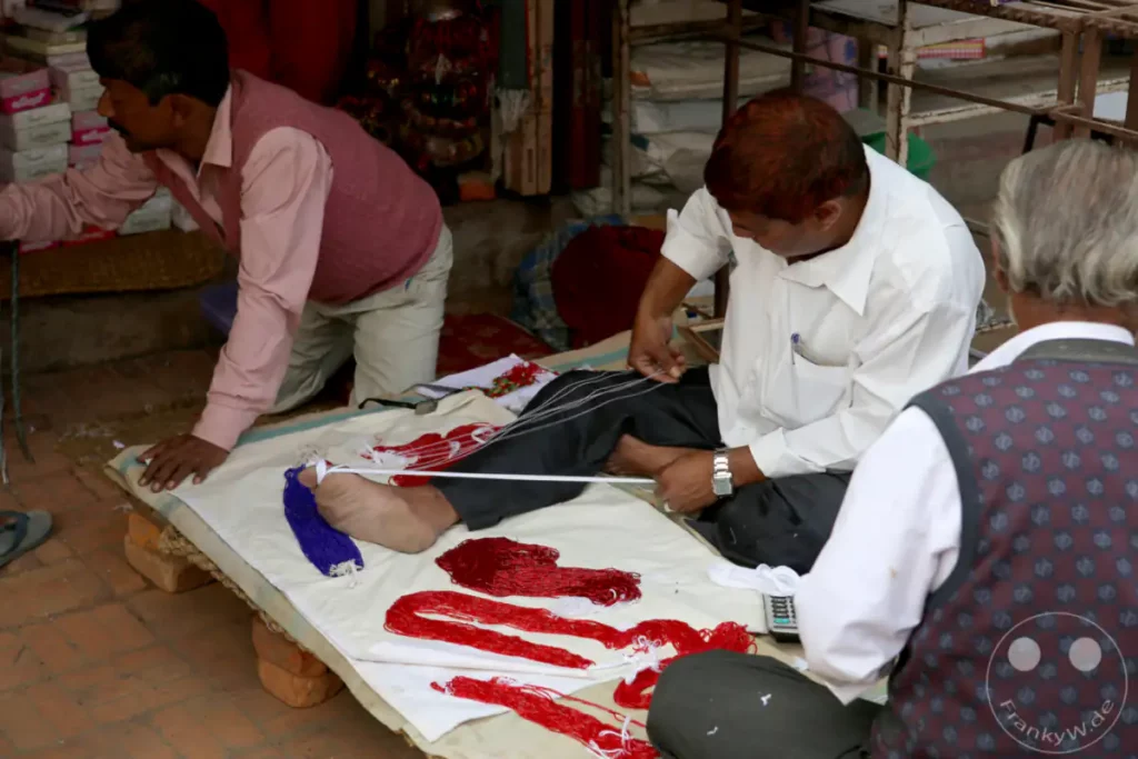 Männer sitzen auf dem Boden und arbeiten an Perlenprodukten, wie es auf dem Durbar Square in Patan üblich ist. Sie sind mit der Herstellung von Perlenketten und anderen Handwerksartikeln beschäftigt, was eine typische Szene auf diesem historischen Platz darstellt.