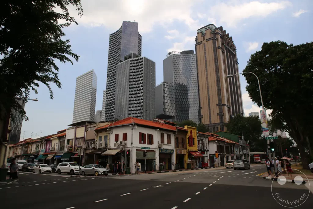 Eine Straße in Singapur, die von traditionellen Gebäuden mit bunten Fassaden und modernen Hochhäusern im Hintergrund geprägt ist. Auf der Straße bewegen sich Autos und Fußgänger, was auf ein lebhaftes städtisches Umfeld hindeutet. Die Szene kombiniert historische Architektur mit zeitgenössischer Stadtentwicklung unter einem bewölkten Himmel.
