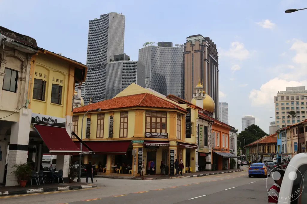 Eine Straße in Singapur, die traditionelle zweistöckige Gebäude mit modernen Hochhäusern im Hintergrund verbindet. Auf der linken Seite befinden sich Restaurants mit Sitzgelegenheiten im Freien und indischer Küche. Die Szene vermittelt einen lebendigen Eindruck von der Stadt, in der Altes auf Neues trifft.