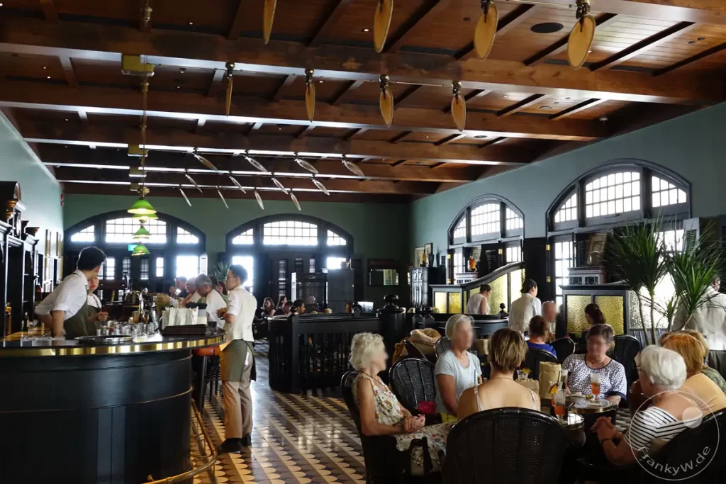 Ein Blick in ein stilvolles Restaurant und Bar-Interieur in Singapur, wo Gäste an Tischen sitzen und sich unterhalten. Die Holzdecke mit dekorativen Elementen und die großen Fenster schaffen eine elegante Atmosphäre. Das Bild vermittelt eine entspannte und gesellige Stimmung während des Tages.