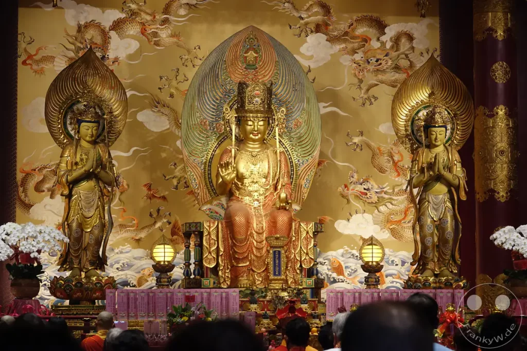 Eine prächtige goldene Buddha-Statue steht im Zentrum eines reich verzierten Tempels, flankiert von zwei kleineren goldenen Statuen und umgeben von Wandmalereien mit Drachenmotiven. Der Altar ist mit Blumen und religiösen Schriftrollen geschmückt, während Gläubige im Vordergrund aus der Ferne zuschauen. Die Szene strahlt eine Atmosphäre von Ehrfurcht und spiritueller Hingabe aus, unterstrichen durch das warme Licht und die goldenen Farben.