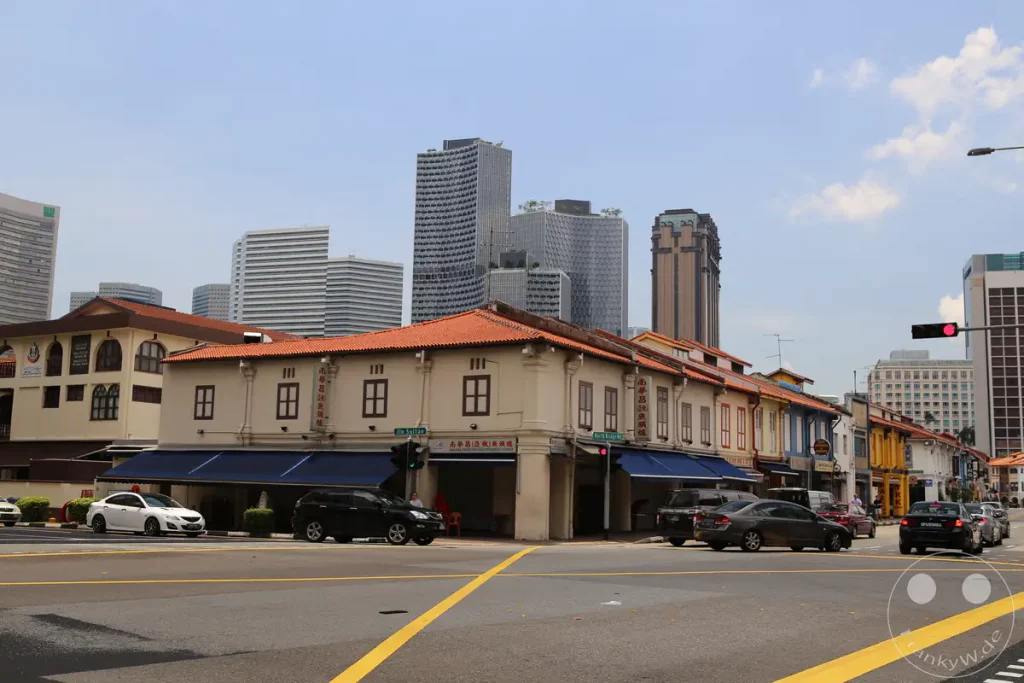 Eine Straße in Singapur zeigt eine Mischung aus historischen, niedrigen Gebäuden mit orangefarbenen Ziegeldächern und imposanten modernen Wolkenkratzern im Hintergrund. Mehrere Autos bewegen sich auf der Straße, während das Tageslicht und der blaue Himmel eine geschäftige, aber klare Atmosphäre schaffen.