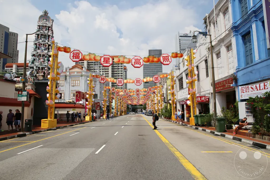 Eine Straße in Chinatown, Singapur, ist mit zahlreichen roten und gelben Laternen geschmückt, die für ein Fest oder eine Feierlichkeit aufgehängt wurden. Die Gebäude im Hintergrund sind im typischen Stil der Region gehalten, während auf dem Gehweg Menschen flanieren und die festliche Atmosphäre genießen. Moderne Hochhäuser bilden einen Kontrast zur traditionellen Architektur und den Dekorationen.