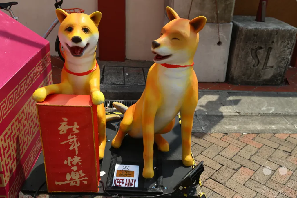 Zwei dekorative Hundestatuen stehen vor einem roten Behälter, der mit chinesischen Schriftzeichen verziert ist, und einer pinken Kiste mit einem geometrischen Muster. Eine der Statuen liegt, während die andere aufrecht sitzt und eine rote Schleife um den Hals trägt. Im Hintergrund sind ein Fahrrad und ein Betonsockel mit der Aufschrift "SL" zu sehen, was auf eine öffentliche Dekoration in Singapur hindeutet.