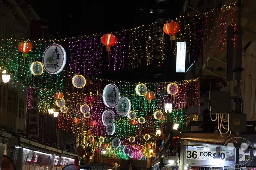 Bunte Lichterketten und traditionelle rote Laternen schmücken die Straßen von Chinatown in Singapur bei Nacht. Kreisförmige Dekorationen mit chinesischen Motiven hängen zwischen den Lichtern und erzeugen eine festliche Atmosphäre. Die belebte Gasse lädt mit ihren Geschäften und Lichtern zum Bummeln ein.