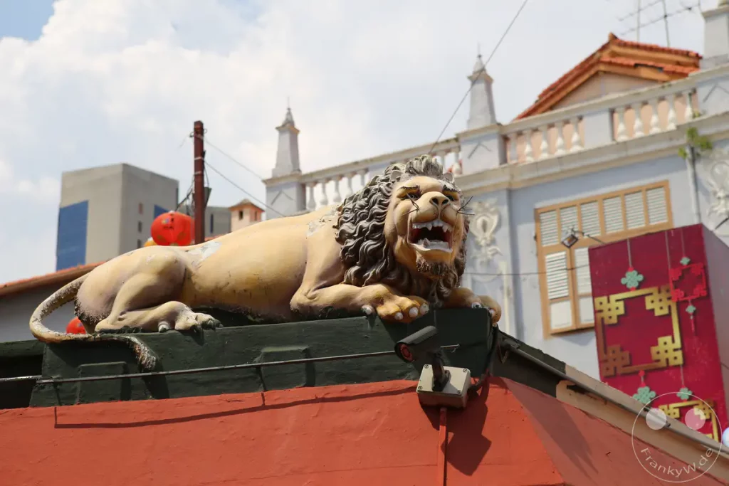 Eine verwitterte Löwenstatue bewacht eine Fassade in Singapurs Chinatown. Im Hintergrund ist eine moderne Hochhausarchitektur zu sehen, die einen starken Kontrast zur traditionellen Umgebung bildet. Die Szene wird von einer Überwachungskamera ergänzt, die über dem Eingangsbereich angebracht ist.