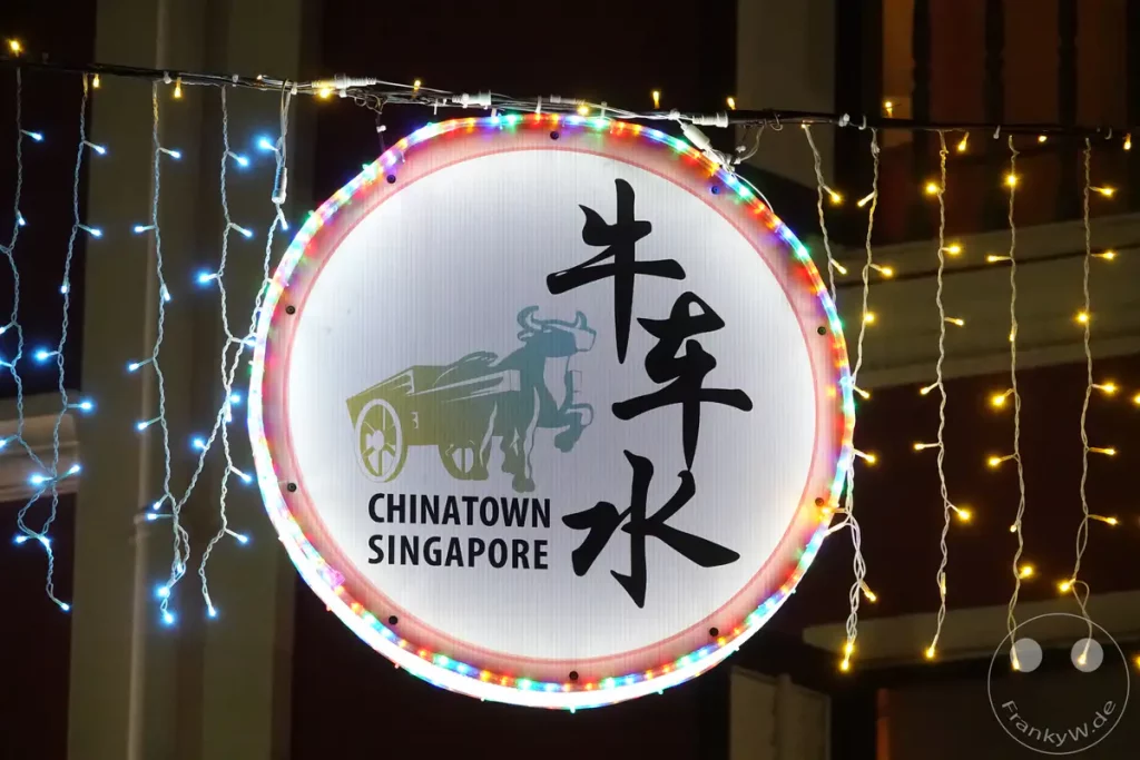 Ein beleuchtetes Schild mit chinesischen Schriftzeichen und der Aufschrift "CHINATOWN SINGAPORE" hängt im Dunkeln. Bunte und weiße Lichterketten zieren das Schild und die Umgebung, was eine festliche und einladende Atmosphäre schafft. Das Bild fängt die nächtliche Belebung eines belebten Viertels ein.
