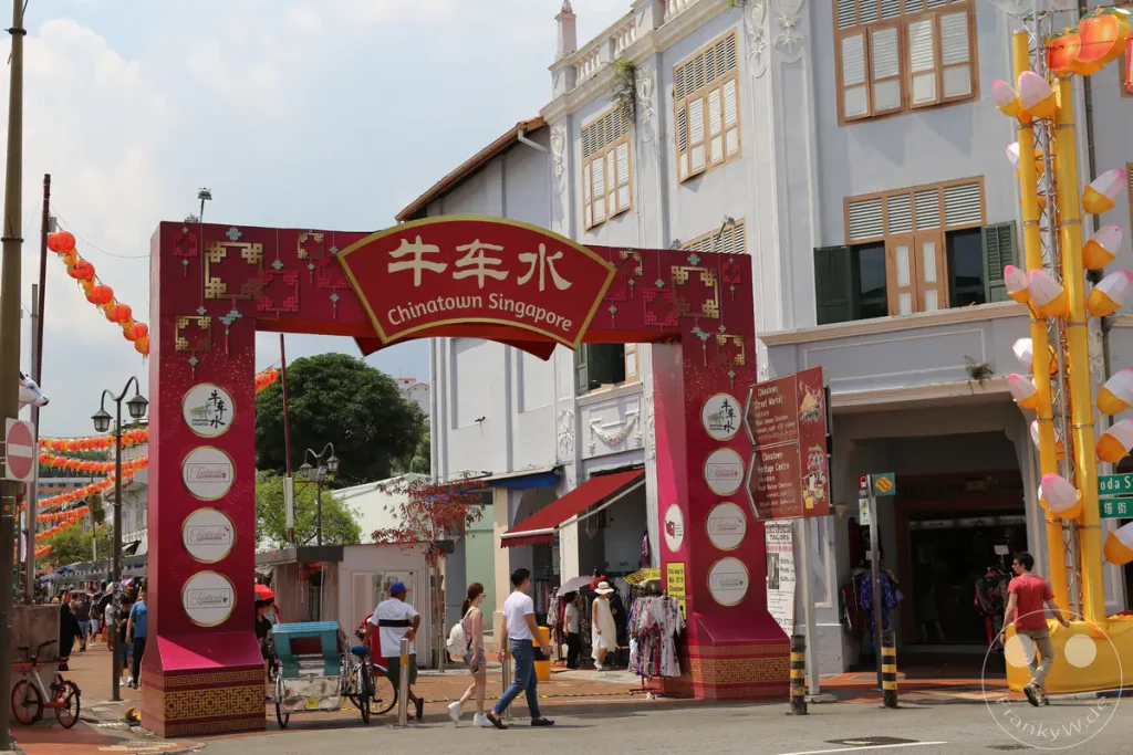 Ein farbenfroher Torbogen markiert den Eingang zu Chinatown in Singapur, geschmückt mit chinesischen Schriftzeichen und traditionellen Mustern. Entlang der Straße hängen orange und rote Laternen, die eine festliche Atmosphäre schaffen. Menschen schlendern durch die belebte Straße, vorbei an Geschäften und Ständen, die lokale Waren anbieten.