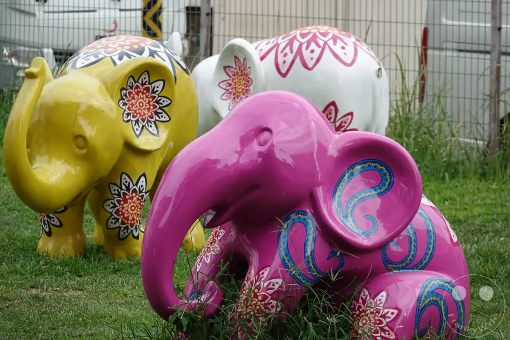Mehrere bunte Dekorationselefanten stehen auf einer grünen Rasenfläche in Singapur. Die Elefanten sind mit floralen Mustern und Paisleymotiven verziert. Die Szenerie wirkt verspielt und künstlerisch.