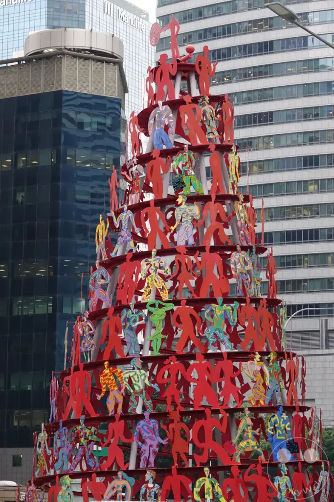 Eine dynamische Skulptur aus roten, abstrakt dargestellten menschlichen Figuren dominiert den Vordergrund des Bildes. Sie befindet sich vor der modernen Glasfassade eines Hochhauses in Singapurs Innenstadt. Die bunten Muster auf einigen Figuren und die Spiegelungen in den Fenstern erzeugen eine lebendige und urbane Atmosphäre.