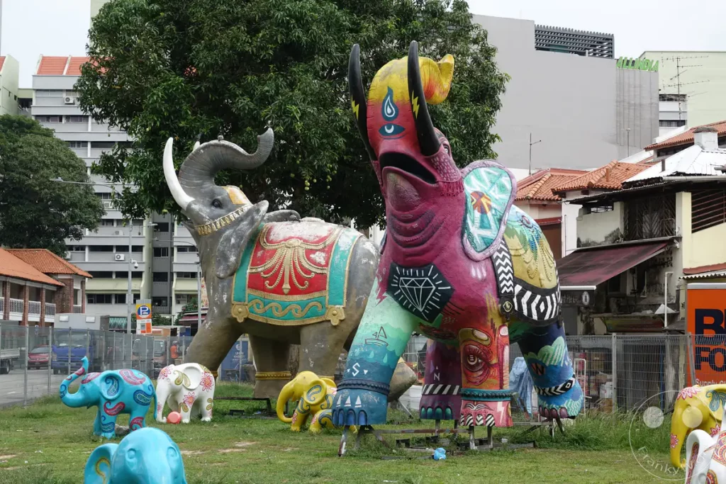 Bunte Skulpturen von Elefanten stehen auf einer Rasenfläche in Singapur. Die Kunstwerke sind mit leuchtenden Farben und Mustern verziert, darunter auch asiatisch inspirierte Paisleymotive. Im Hintergrund sind Gebäude und Bäume zu sehen, die auf eine städtische Umgebung hinweisen.
