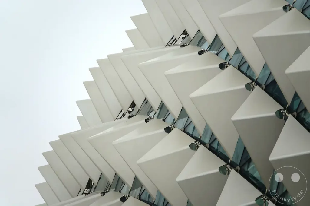 Die markante, stachelartige Fassade der Esplanade in Singapur präsentiert sich unter einem bewölkten Himmel. Geometrische Elemente aus weißen Paneelen überlappen sich und erzeugen ein dynamisches Muster. Die Struktur wirkt modern und erinnert an eine Muschel oder einen Fächer.