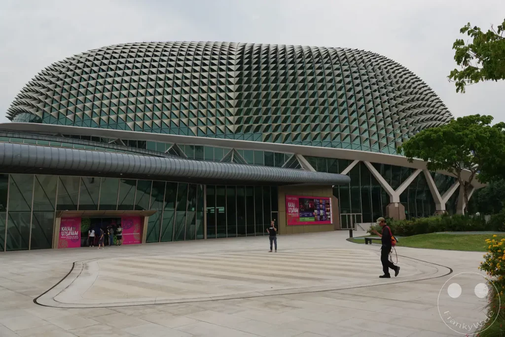 Die markante, säbel- oder durian-förmige Architektur der Esplanade – Theatres on the Bay in Singapur dominiert das Bild. Die Fassade besteht aus einer komplexen Struktur von Aluminiumpaneelen, die sich wie eine Haut über das Gebäude spannen. Im Vordergrund bewegt sich eine Person über den gepflasterten Platz vor dem Gebäude, während eine üppige grüne Baumkrone die Szene im Hintergrund ergänzt.