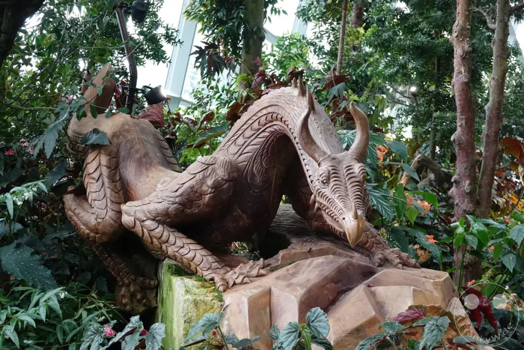 Eine kunstvolle Drachenskulptur aus Holz ruht inmitten einer üppigen tropischen Pflanzenpracht im "Gardens by the Bay" in Singapur. Das detailreiche Kunstwerk fügt sich harmonisch in die natürliche Umgebung ein und erzeugt eine mystische Atmosphäre. Zahlreiche grüne und rötliche Blätter sowie zarte Blüten umrahmen die Skulptur und vermitteln das Gefühl, sich in einem geheimen Garten zu befinden.