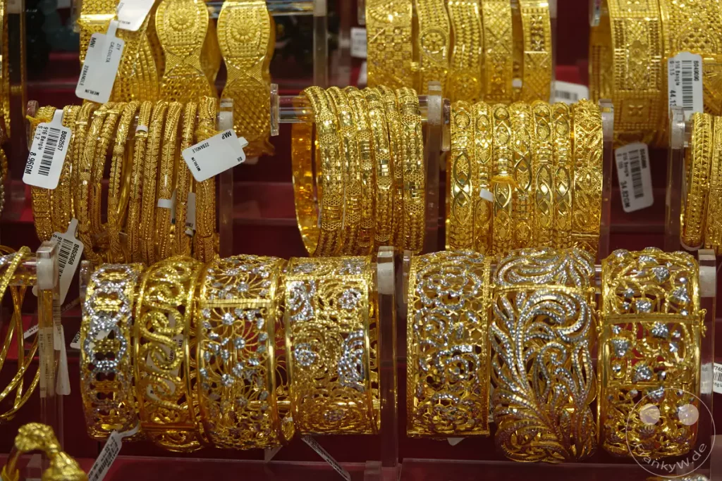 Eine Nahaufnahme von kunstvoll gefertigten goldenen Armreifen, die in einem Geschäft in Singapur ausgestellt sind. Die Vielfalt der Muster und die glänzende Oberfläche des Goldes vermitteln ein Gefühl von Reichtum und Eleganz. Die traditionellen Designs deuten auf kulturelle Bedeutung und feine Handwerkskunst hin.