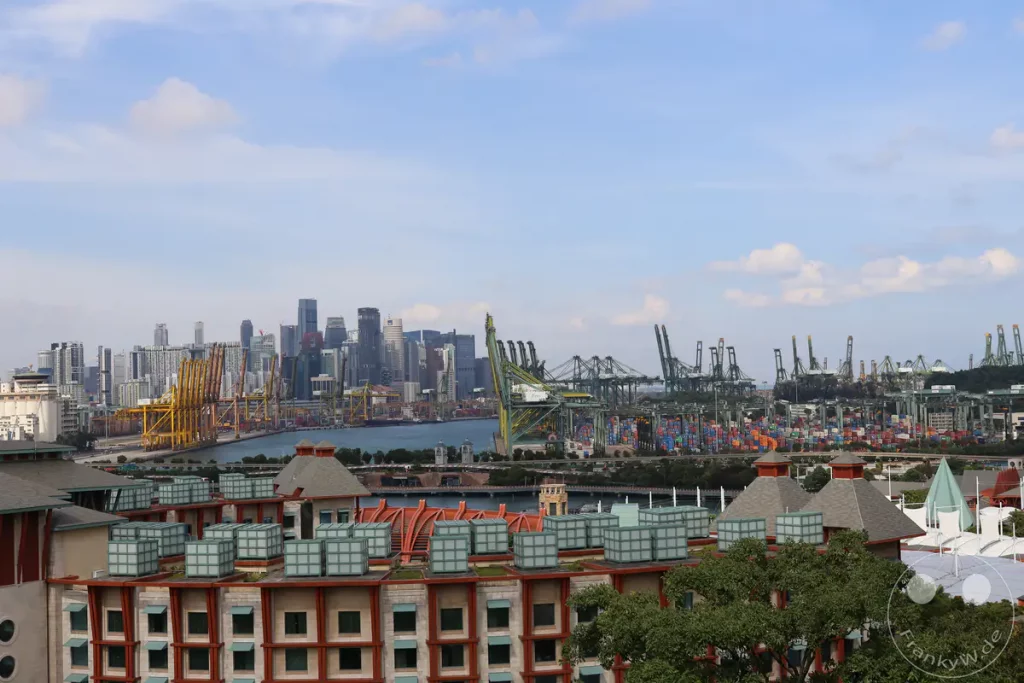 Das Bild zeigt eine weitläufige Hafenszenerie in Singapur mit zahlreichen Containerterminals und großen Kränen im Vordergrund. Im Hintergrund erhebt sich die beeindruckende Skyline der Stadt mit ihren modernen Hochhäusern. Die Aufnahme vermittelt einen Eindruck von geschäftiger Betriebsamkeit und urbaner Dynamik.