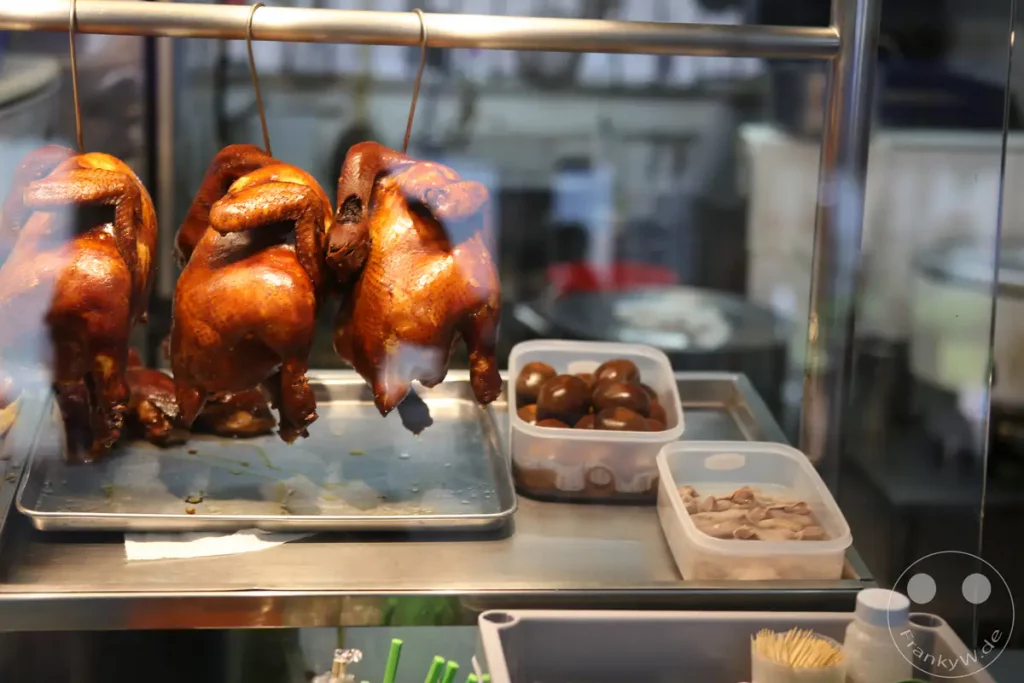 Mehrere saftig aussehende, geröstete Hähnchen hängen an Haken über einer Metallablage, mit weiteren appetitlich aussehenden Speisen daneben. Die Speisen sind hinter Glas in einer Auslage eines Hawker-Centers in Singapur präsentiert. Das Ambiente wirkt lebendig und kulinarisch vielfältig.
