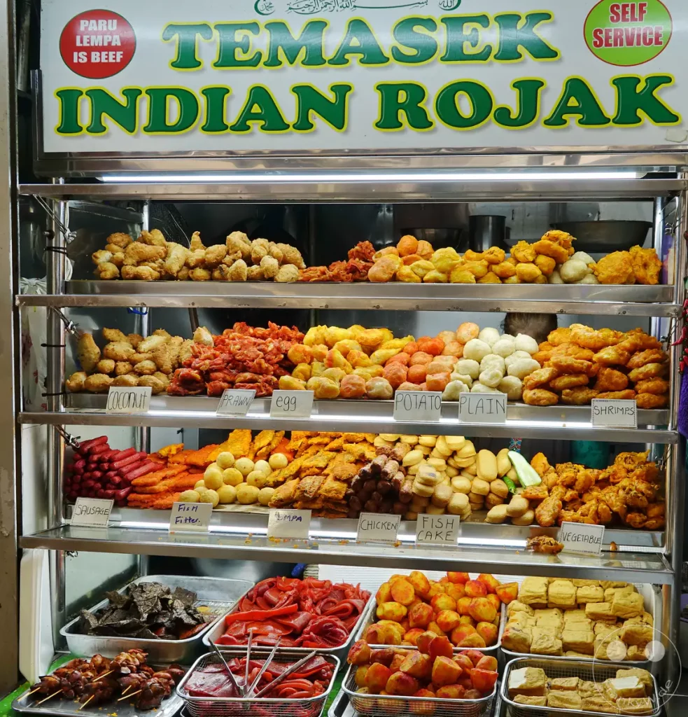 Einladender Essensstand in Singapur mit einer Vielfalt an frittierten indischen Spezialitäten. Die Auslage ist auf mehreren Ebenen präsentiert und lädt zur Selbstbedienung ein. Die bunten Speisen und die klare Beschilderung erzeugen eine lebendige und appetitliche Atmosphäre.