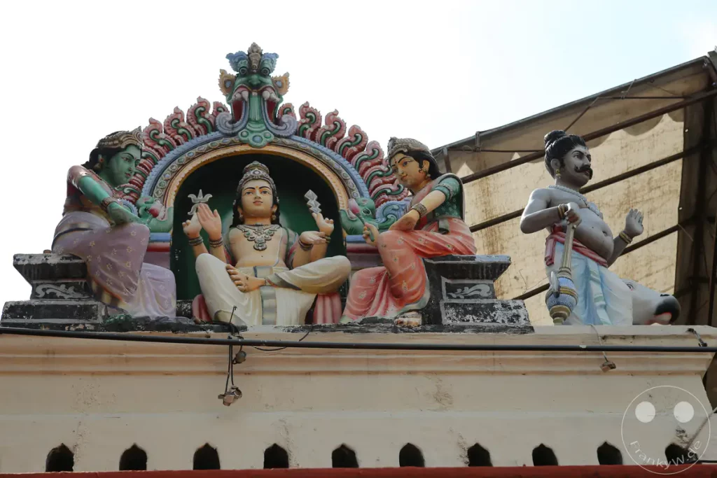 Mehrere bunte Skulpturen von hinduistischen Gottheiten zieren die Fassade eines Tempels in Singapur. Die Figuren, dargestellt mit traditionellen Gewändern und Schmuck, sitzen und stehen auf einem verzierten Sockel unter einem Sonnenschutz. Die aufwendige Architektur und die lebendigen Farben schaffen eine festliche und spirituelle Atmosphäre.